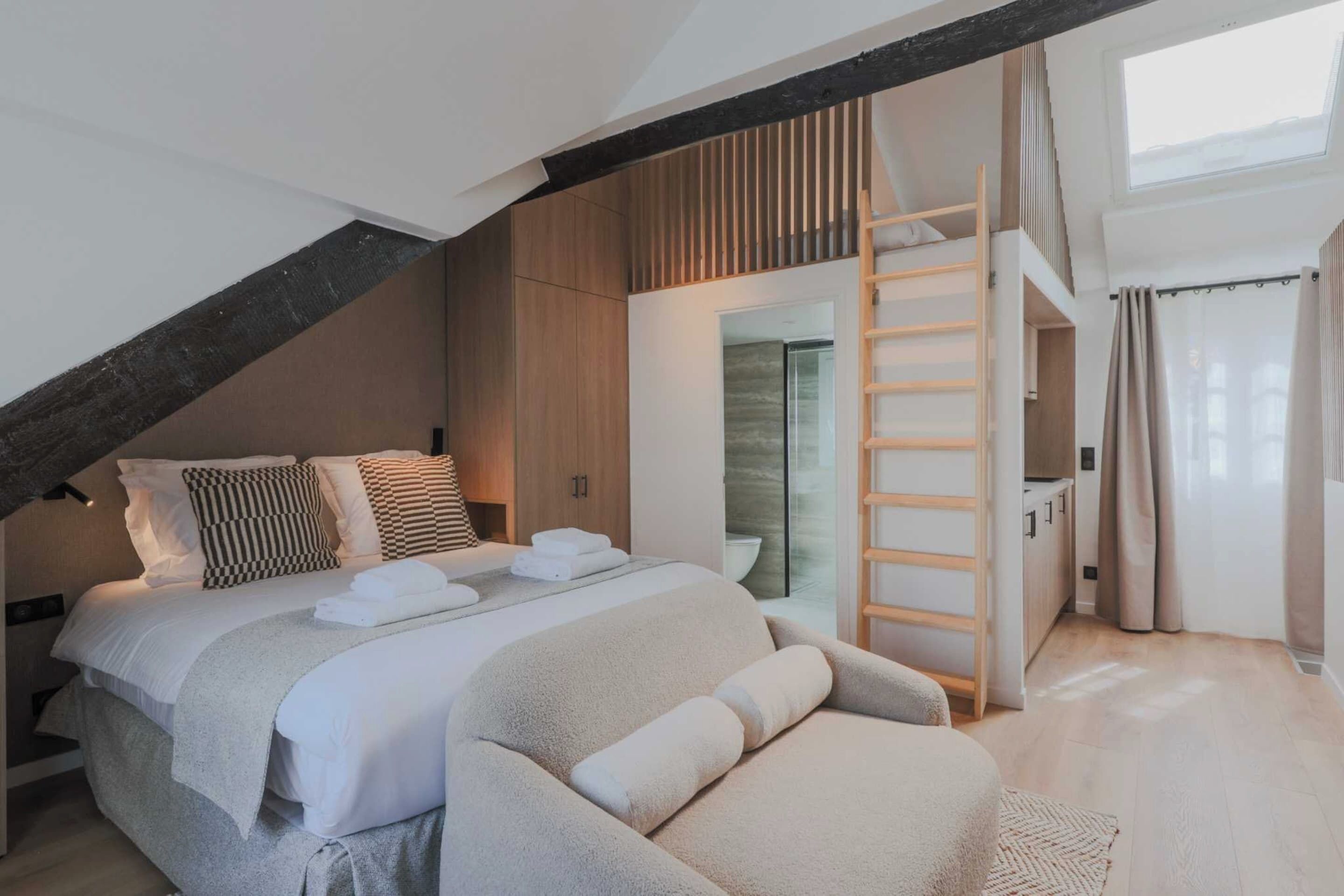 A bedroom with a lofted sleeping area, a wooden ladder, and a bathroom with a glass door.