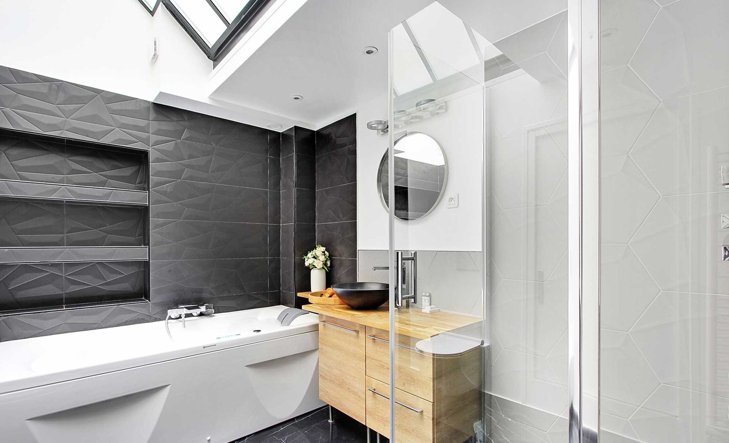 A modern bathroom with a freestanding bathtub, a wooden vanity, a round mirror, and a geometric-patterned wall.
