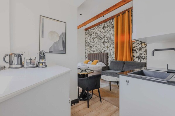 Habitación de hotel compacta con kitchenette, cama con cabecera gris, sofá gris y mesa de comedor.