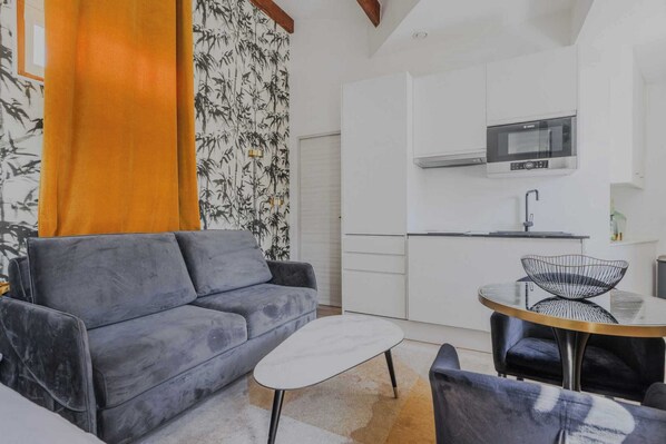 Ein kompakter Wohnbereich mit grauem Sofa, einem kleinen runden Tisch und einer Küchenzeile inklusive Mikrowelle und Schränken.