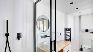 A modern bathroom with a wooden vanity, a round mirror, a towel rack, a toilet, and a washing machine. - Charming Studio A/C - 2P - Le Marais (Paris)
