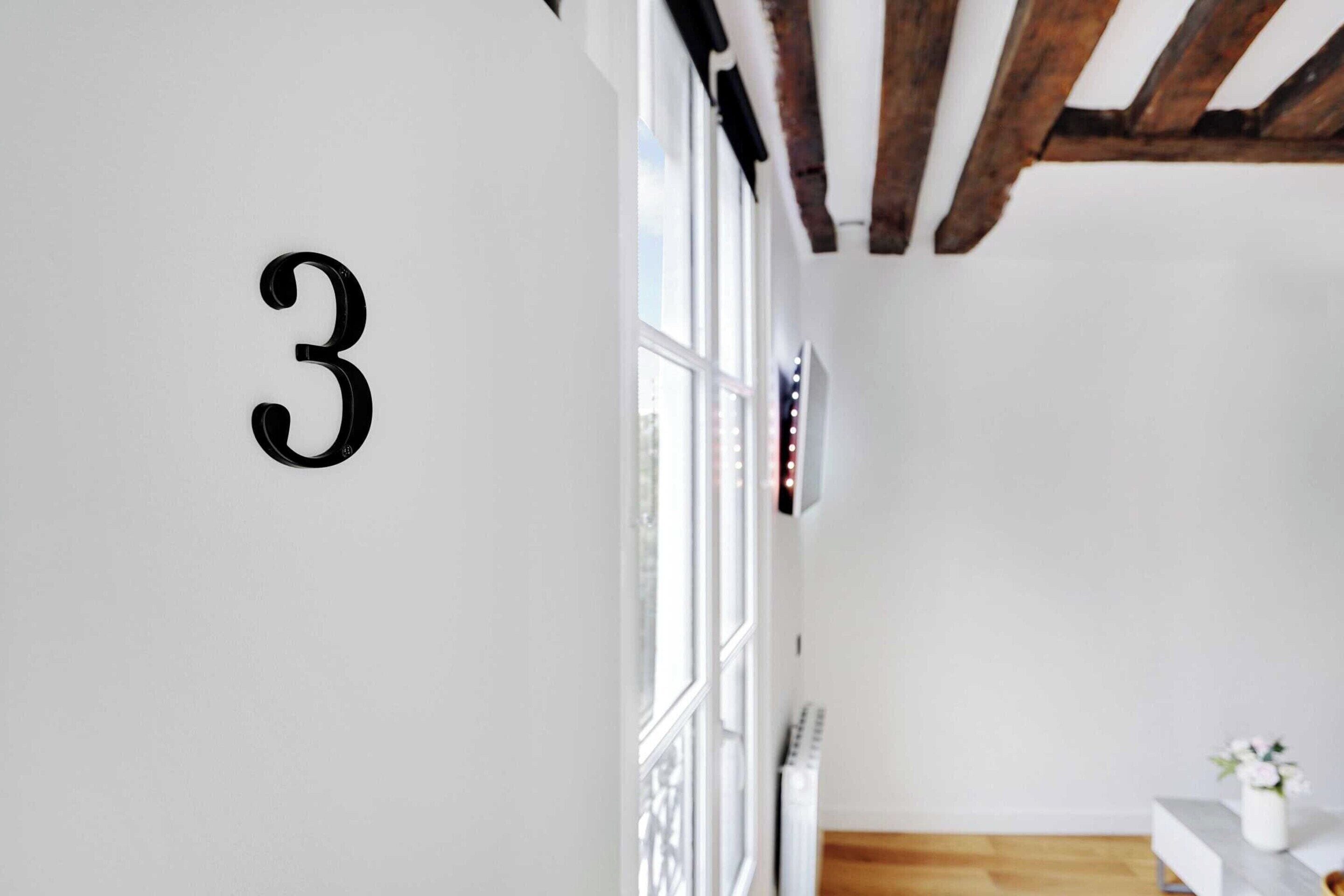 A room with a large number '3' on the wall, a window with blinds, a radiator, and a small table with a vase of flowers.