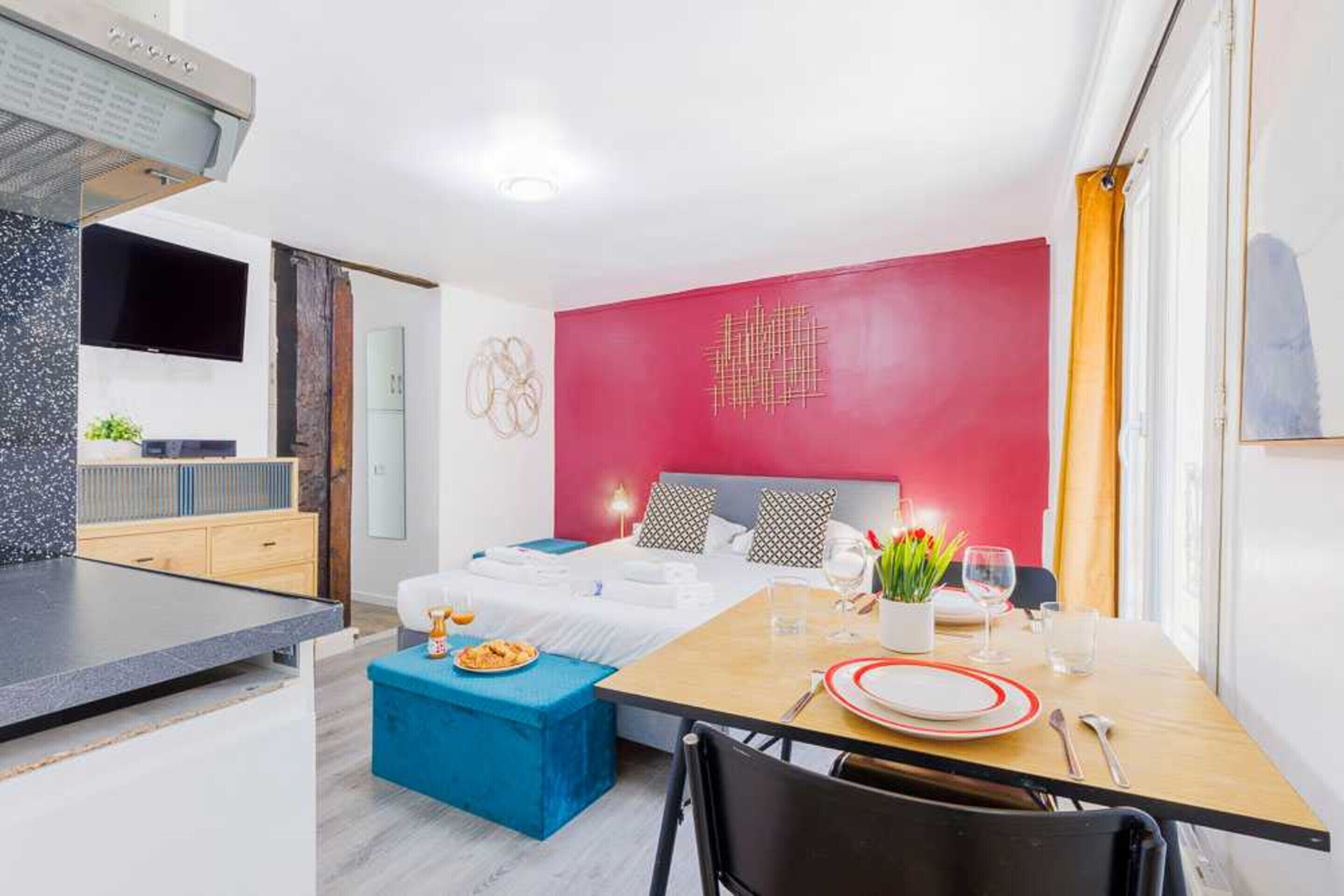 A modern hotel room with a bed, a dining table, a kitchen area, and a red accent wall.