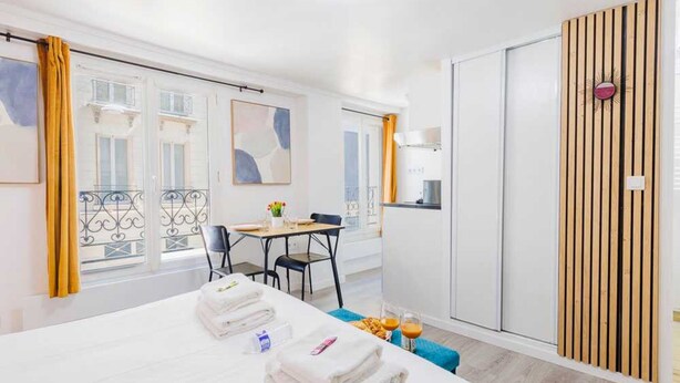 Habitación de hotel con cama, mesa y sillas, una pequeña cocina y un balcón con cortinas amarillas.