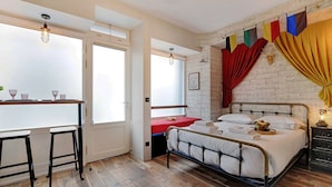 A bedroom with a bed, a desk with stools, a window, and a brick wall. - The Wizard's Studio! (Paris)
