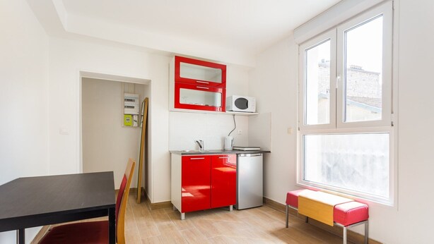 Une cuisine compacte avec un meuble rouge, un four à micro-ondes et un petit évier. On y trouve une table à manger avec une chaise et un banc près de la fenêtre.