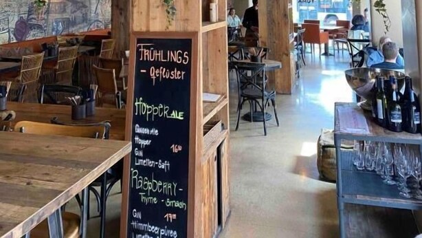 Un restaurant chaleureux avec des tables et des chaises en bois, un menu écrit à la craie et des plantes suspendues.