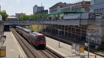 Un andén de estación de tren con un tren sobre las vías, un puente peatonal y edificios al fondo.
