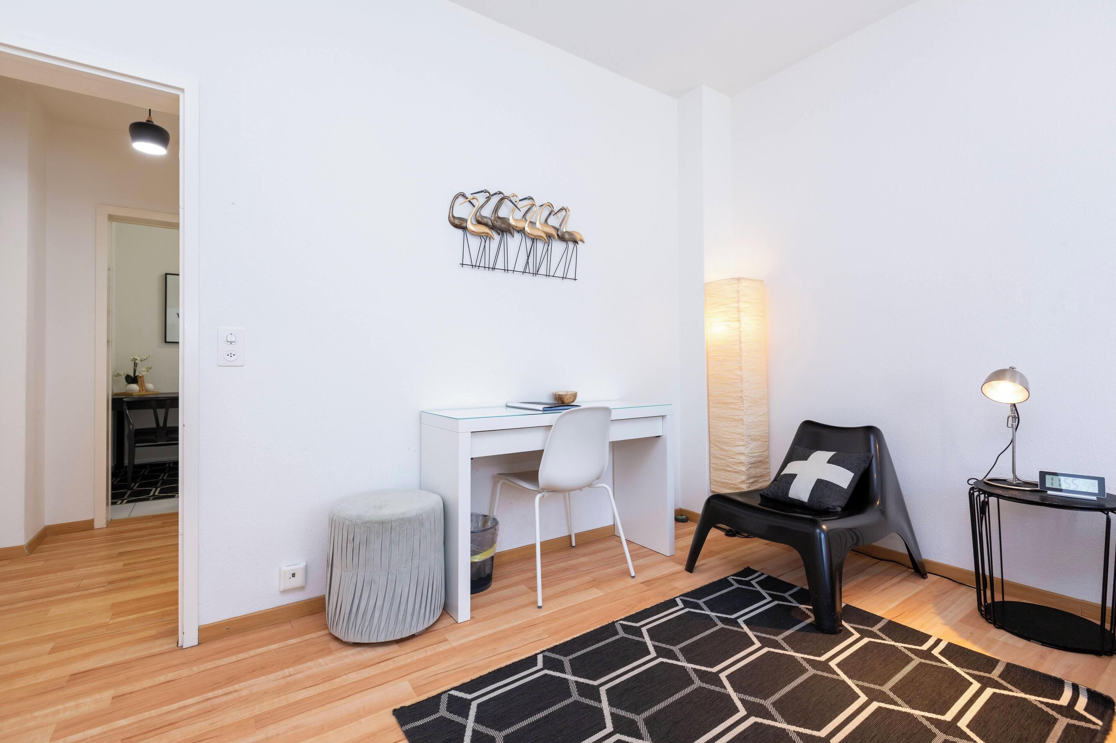 Una habitación moderna y bien iluminada, con un escritorio blanco, una silla negra, una mesita auxiliar pequeña y una alfombra con estampado geométrico.