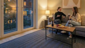 Un salon confortable avec un canapé, une table basse sur laquelle sont disposés des en-cas et une bouteille de vin, et de grandes fenêtres offrant une vue sur un immeuble et des arbres.