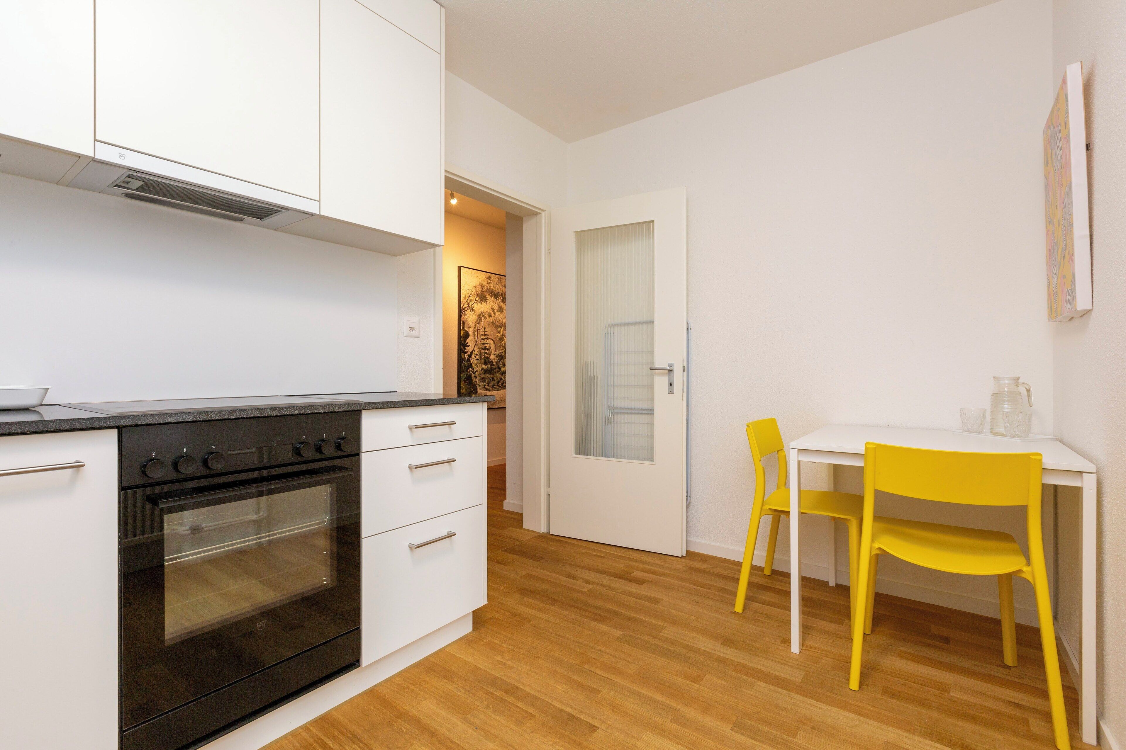 Une cuisine avec des meubles blancs, un four noir et un plancher en bois. On y trouve une table blanche, une chaise jaune et une petite fenêtre.