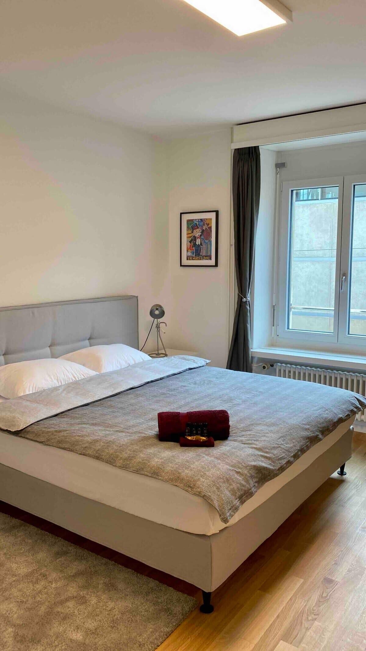 A neatly arranged bedroom with a bed, a bedside lamp, a framed picture on the wall, and a window with curtains.
