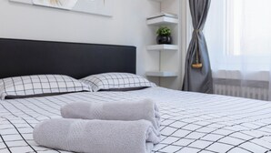 Un lit bien fait, avec une tête de lit noire, une couette à carreaux noirs et blancs et deux serviettes pliées. Une étagère fixée au mur contient des livres et une petite plante.