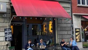 Un espace extérieur aménagé avec des sièges, surmonté d’un auvent rouge, devant un bâtiment en briques, et agrémenté d’une enseigne dans une langue étrangère.