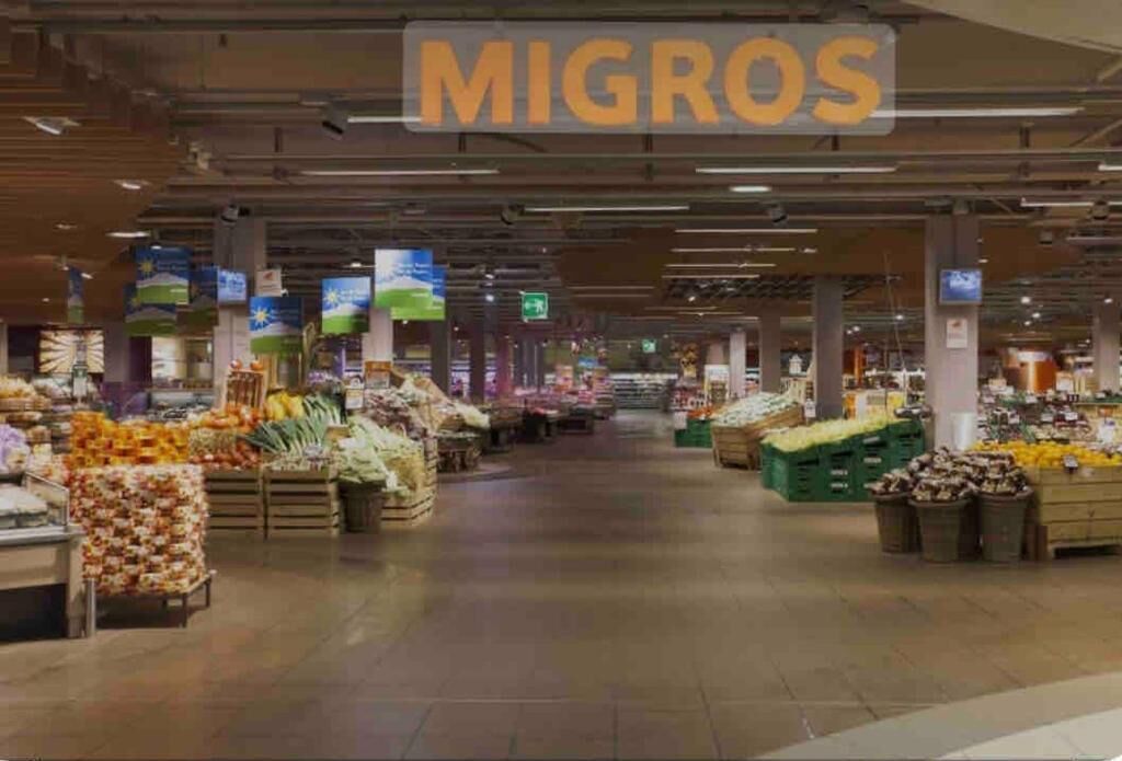 Interieur van een supermarkt met verschillende groente- en fruitafdelingen en een bord met de tekst 'MIGROS'.