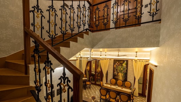 A staircase with ornate black and gold railings leading to a dining area with a table and chairs, and a ceiling fan above. - Ishaan Holiday Home (Alibag)