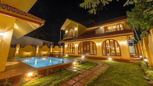 A well-lit, two-story house with a swimming pool, surrounded by a garden and a fence. - Ishaan Holiday Home (Alibag)