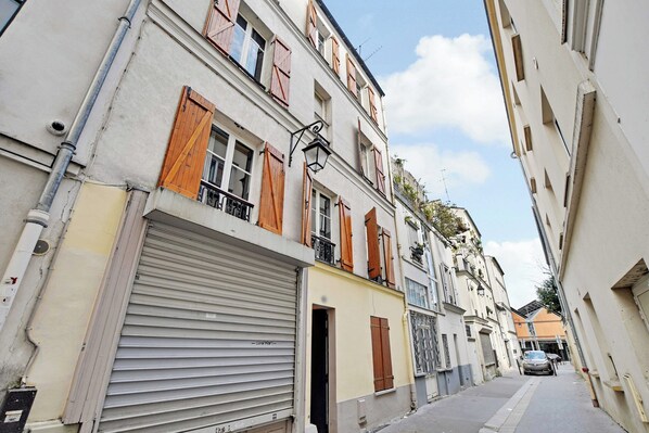 Una calle estrecha flanqueada por edificios, con contraventanas cerradas y la entrada visible a un local en la planta baja.