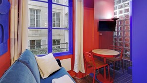 Een kamer met een blauwe bank, een klein rond tafeltje met rode stoelen, een aan een muur bevestigde televisie en een raam met blauwe kozijnen.