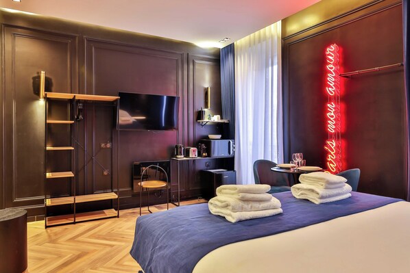 A modern hotel room with a large bed, a flat-screen TV, a desk with a chair, and a neon sign on the wall.