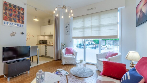 A modern living room with a flat-screen TV, a coffee table, and a sofa with red and blue cushions. There is a dining area with a white chair and a small table. The room has large windows with a view of the outside. - Cosy Studio - 2P - Port de Deauville (Deauville)