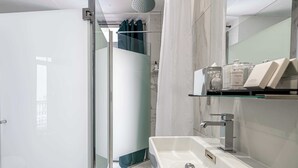 A modern bathroom with a glass shower enclosure, a white sink, and a mirror with toiletries. - Hotel Avaton - Deluxe Balcony Bedroom - Cannes Palais (Cannes)