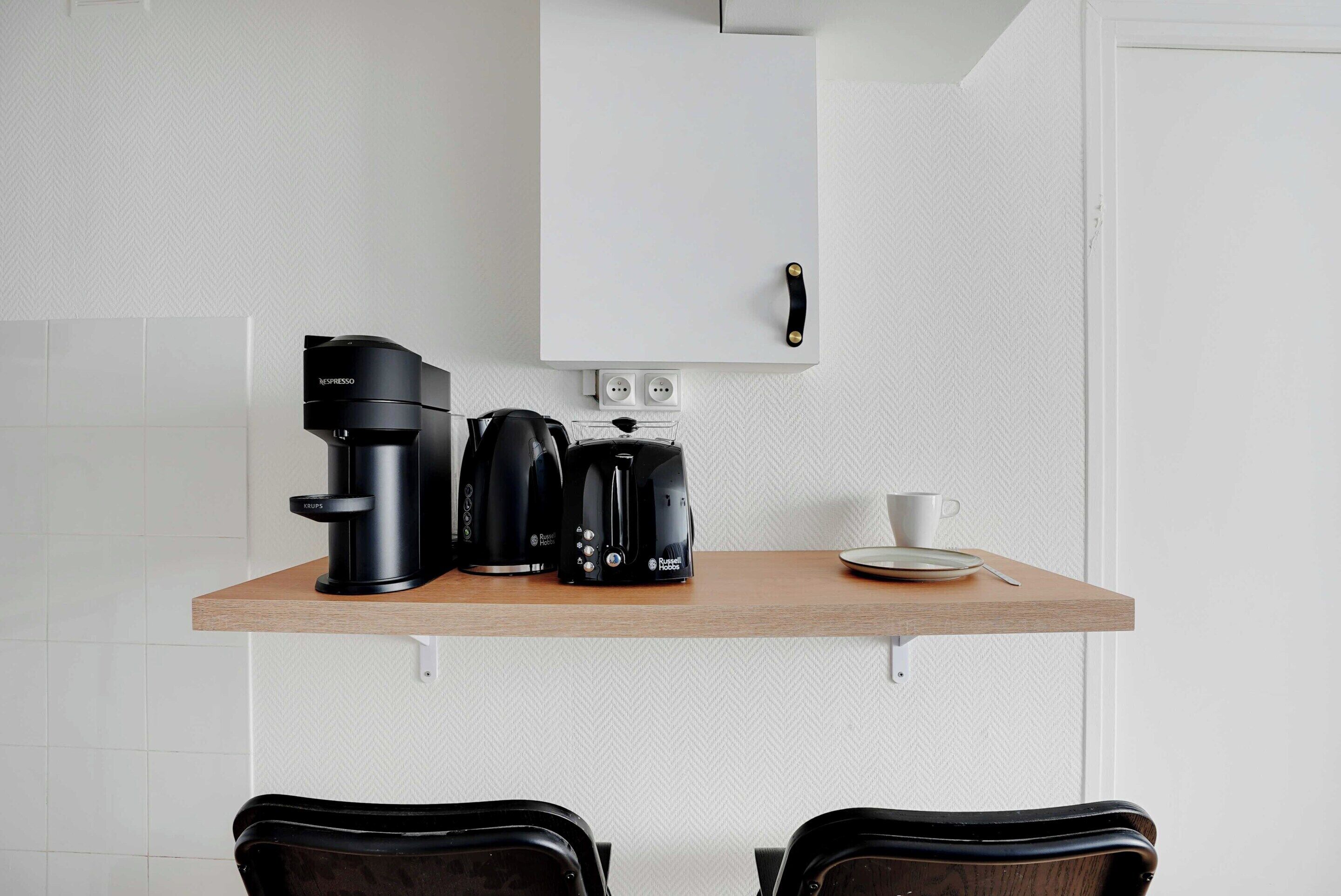 Une petite cuisine équipée d’une machine à café, d’une bouilloire et d’une tasse, le tout posé sur un plan de travail en bois.
