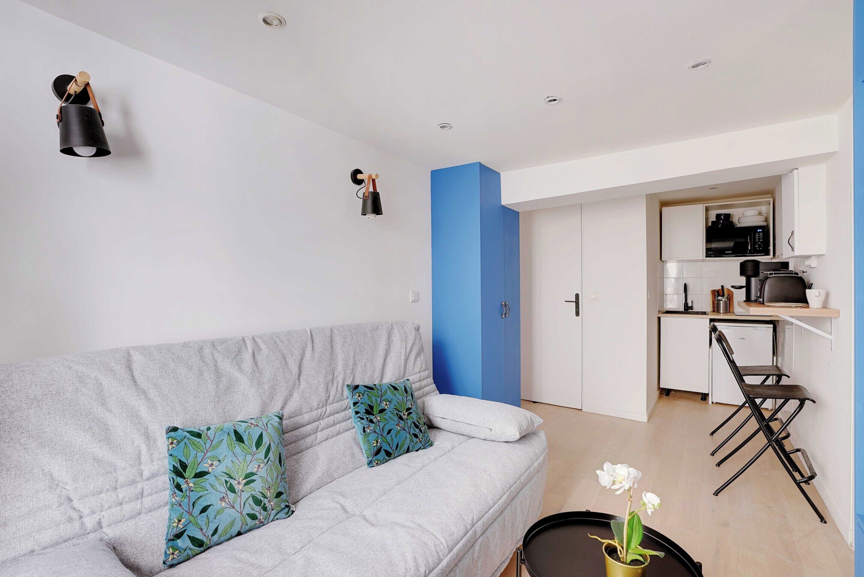 Una sala de estar moderna con un sofá gris, una mesita redonda con una planta y una cocina al fondo.