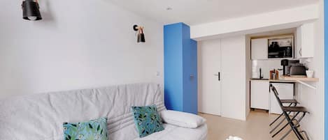 Una sala de estar moderna con un sofá gris, una mesita redonda con una planta y una cocina al fondo.