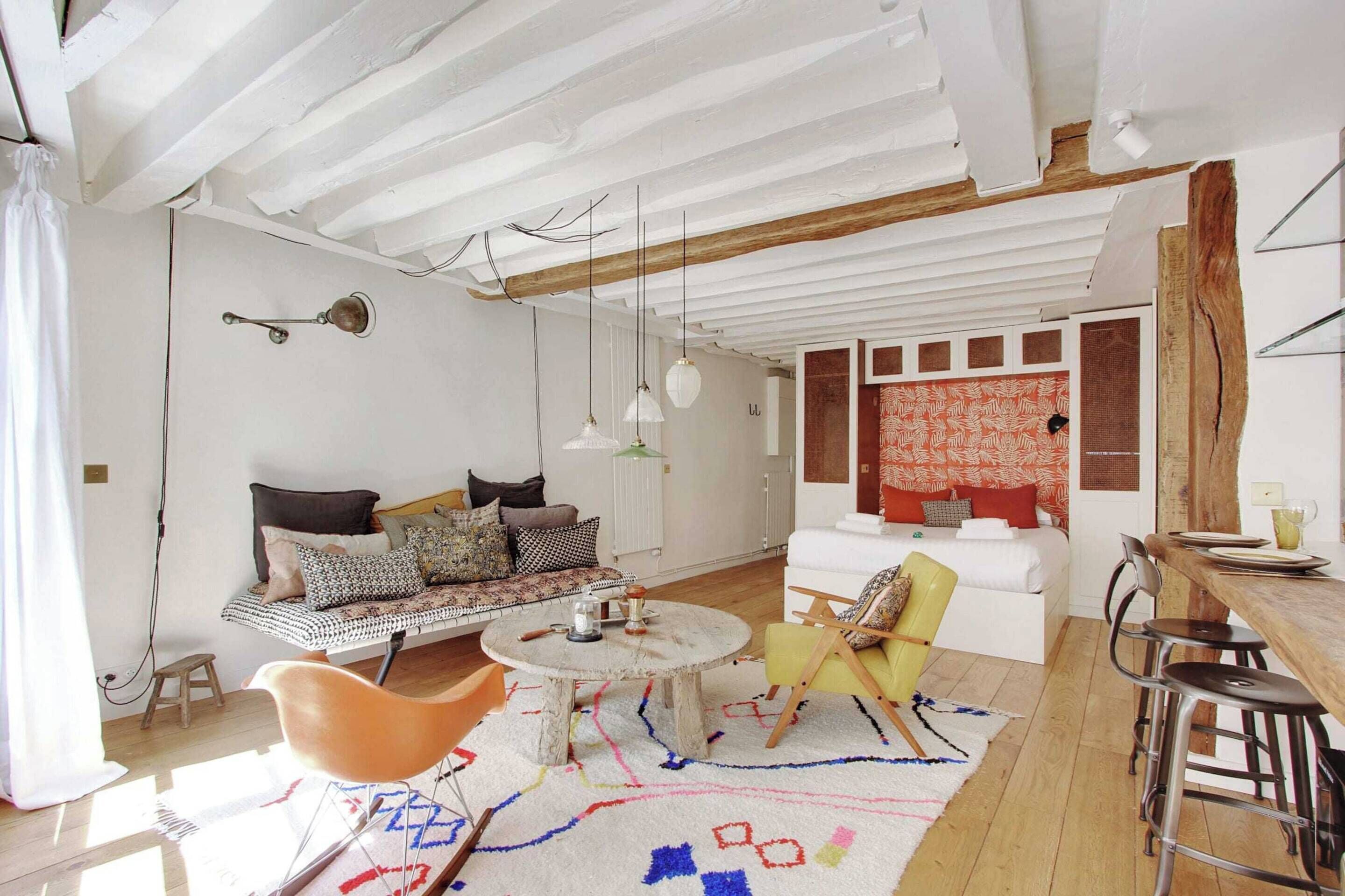 A cozy living room with a wooden ceiling, a round coffee table, a sofa, a yellow armchair, and a dining area with a wooden table and bar stools.