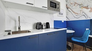 A modern kitchen with a built-in microwave, a coffee maker, and a sink. There is a blue chair and a small table in the adjacent dining area. - Modern Sutdio for 2 With A/C - Near Montmartre (Paris)