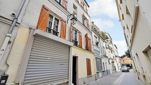 A narrow street with buildings on either side, featuring closed shutters and a visible entrance to a ground-floor establishment. - Modern Sutdio for 2 With A/C - Near Montmartre (Paris)