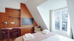 A modern hotel room with a sloped ceiling, a bed with white linens, a desk with two purple chairs, a TV mounted on the wall, and a window with a view of the city. - Elegant Studio - 2P - Republique (Paris)