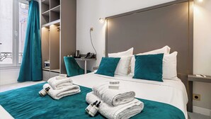 A modern hotel room with a large bed, teal bedding, a built-in wardrobe, a bedside table with a lamp, and a window with curtains. - Avaton Hotel - Deluxe Room - Cannes Palais (Cannes)