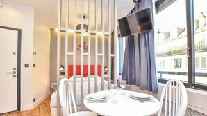 Una habitación de hotel moderna con zona de comedor que incluye una mesa redonda, sillas blancas y vistas a la ciudad a través de amplios ventanales.