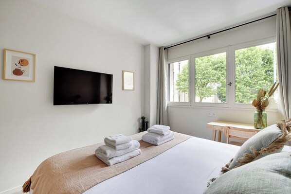 A hotel room with a bed, a TV mounted on the wall, a desk, and a window with a view of trees. - Modern "green Suites" Residence - Meudon/velizy (Meudon)