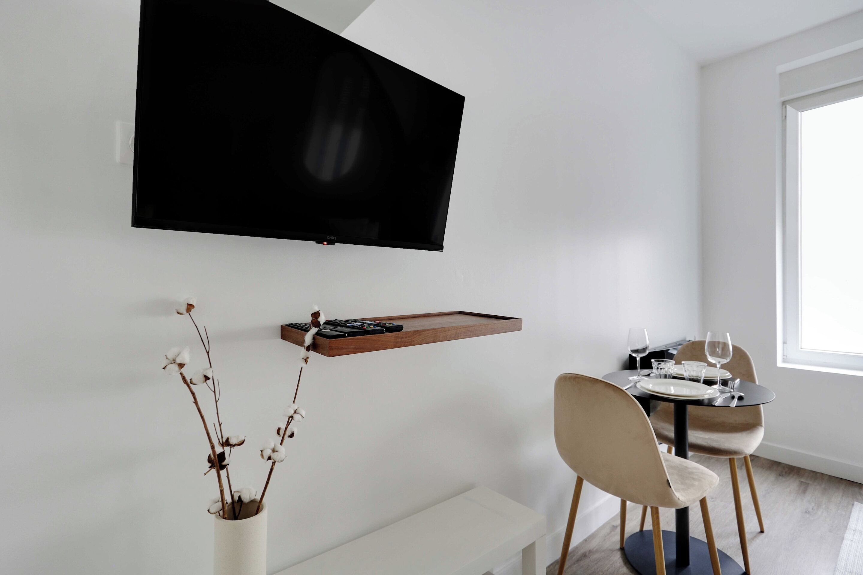 Un'area pranzo moderna con una TV a schermo piatto montata a parete, una mensola in legno con libri e un vassoio, un vaso con rami secchi e un tavolo da pranzo apparecchiato con piatti e bicchieri.