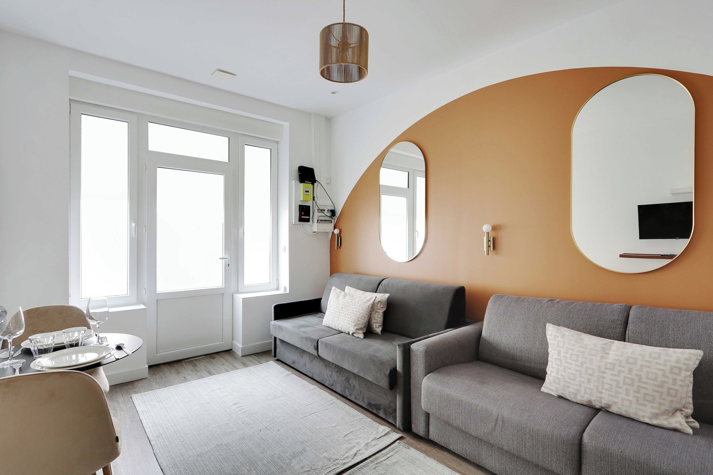 Un salon moderne avec un canapé gris, un miroir rond et un coin repas avec une table dressée pour quatre personnes.