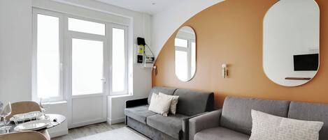 Un salon moderne avec un canapé gris, un miroir rond et un coin repas avec une table dressée pour quatre personnes.