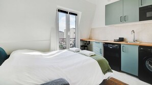 Une chambre compacte comprenant un lit, une petite kitchenette et une fenêtre donnant sur des immeubles.
