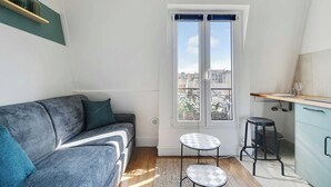 Un espace de vie compact comprenant un canapé, une petite table, un tabouret, une fenêtre donnant sur un paysage agréable et un coin cuisine.