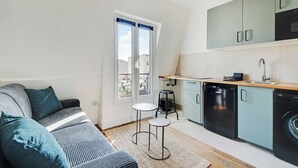 Un espace de vie compact comprenant un coin cuisine, un canapé avec des coussins, une petite table ronde et une fenêtre avec des rideaux.