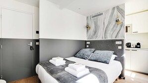 A modern hotel room with a bed, grey and white bedding, a grey headboard, a marble-patterned wall, a small kitchen area, and a grey door. - Cosy Studio for 2P With A/C - Near Montmartre (Paris)