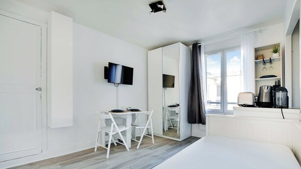 A modern hotel room with a bed, a TV mounted on the wall, a dining table with chairs, a window with curtains, and a kitchenette area. - Cosy Studio - 2P - Place de la Bastille (Paris)