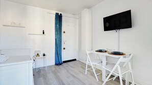 A modern, minimalistic room with a white color scheme, a flat-screen TV mounted on the wall, a small table with chairs, and a door with a blue curtain. - Cosy Studio - 2P - Place de la Bastille (Paris)