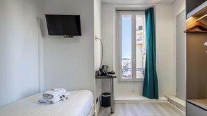A hotel room with a bed, a TV mounted on the wall, a window with curtains, a small table with a trash bin, and a view of buildings outside. - Hotel Avaton - Single Bedroom - Cannes Palais (Cannes)
