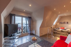 A modern loft bedroom with a large window, a TV on a glass table, and a red sofa. - Apartment With Parking on Place de Morny (Deauville)