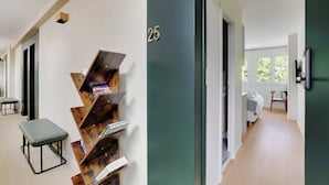 Ein Flur mit einer grünen Tür, die mit der Nummer 25 gekennzeichnet ist, einem Holzregal, einer grünen Bank und einem Blick in ein Schlafzimmer mit Bett und Stuhl.