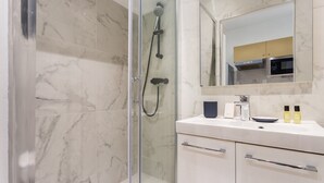 A modern bathroom with a glass-enclosed shower, a white vanity with a mirror, and a kitchen area visible in the reflection. - Cosy Studio - 4P - Palais des Congres (Paris)
