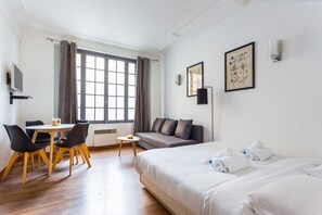 Habitación de hotel con cama, sofá, mesa de comedor con sillas, televisor y obras de arte en las paredes.